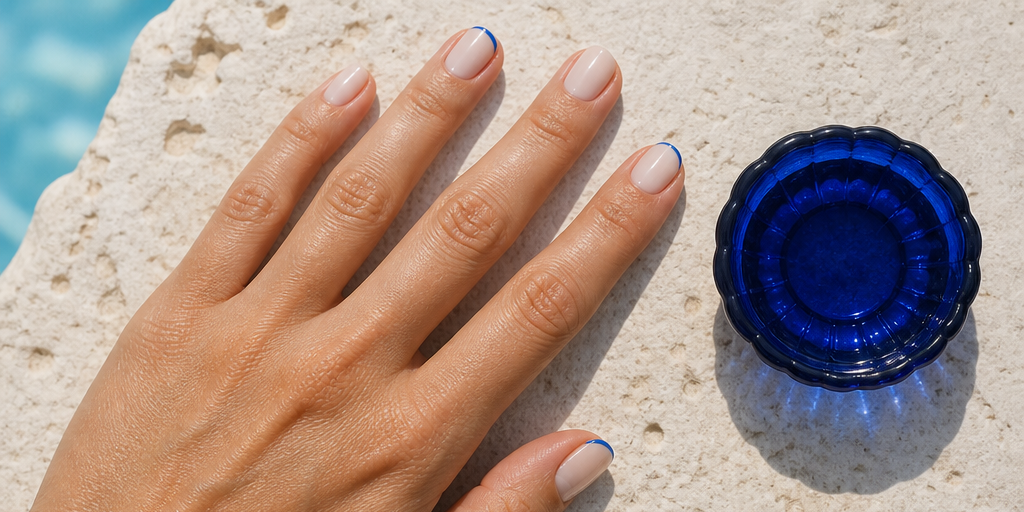 A sheer ivory micro-detail manicure beside Aegean-blue glass on pale stone.